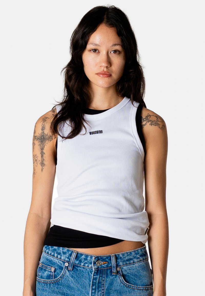 Jeune femme aux longs cheveux foncés portant un débardeur blanc Woodbird par-dessus un t-shirt noir et un jean bleu, montrant des tatouages sur les deux bras supérieurs.