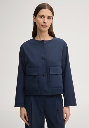 Frau trägt eine marineblaue Jacke mit Knöpfen und großen Klappentaschen, kombiniert mit passenden marineblauen Hosen, steht vor einem schlichten Hintergrund.