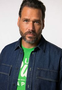 Giacca di jeans con cuciture a contrasto, indossata sopra una maglietta verde con testo bianco. Dettagli in tessuto testurizzato e bottoni visibili.