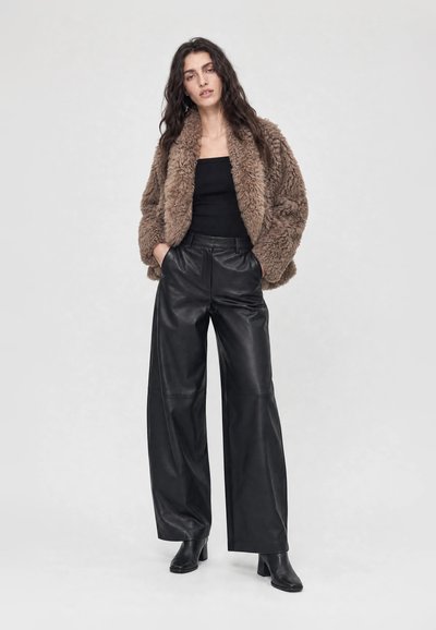 Femme aux longs cheveux foncés portant une veste en fausse fourrure marron, un haut bandeau noir, un pantalon en cuir noir à jambes larges et des bottines noires, debout les mains dans les poches.