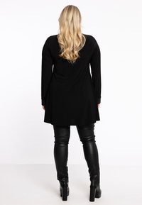 Schwarze Tunika mit langen Ärmeln und fließendem Design, kombiniert mit enganliegenden schwarzen Lederhosen und klobigen Stiefeln mit Absatz. Durchgehend glatte Textur.