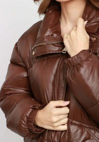Giacca imbottita marrone con finitura lucida, colletto alto e chiusura con zip. Presenta polsini elasticizzati e una sottile trama trapuntata.
