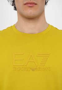Rumena bombažna majica s povečanjem logotipa "EA7 EMPORIO ARMANI" na prsih. Mehka tekstura, okrogla ovratnika, osnovni kroj.