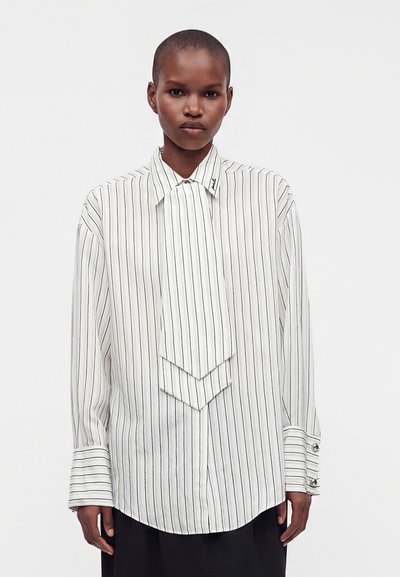 Personne portant une chemise blanche à manches longues rayée de noir verticalement avec un détail de cravate superposée attaché, debout devant un fond uni.