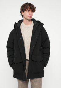 Casaco puffer preto com capuz, fecho frontal de correr e de molas, dois grandes bolsos frontais e um suéter texturizado de malha com padrão de cabo por baixo.