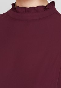 Blusa bordeaux con colletto arricciato, texture liscia e design semplice. Presenta maniche corte e un dettaglio di cucitura sottile.