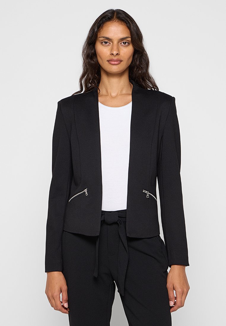 Schwarzer Blazer mit taillierter Passform, schmalen Schultern und zwei Reißverschlusstaschen vorne. Über einem weißen Oberteil getragen, kombiniert mit schwarzen Hosen.