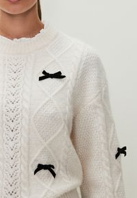 Suéter blanco de punto con un patrón de cables, cuello acanalado y tres pequeños lazos de terciopelo negro como detalles en la parte delantera.