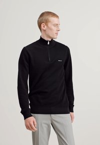 Zwarte zip-up sweater van zacht materiaal, met een hoge kraag en een aansluitend ontwerp. De voorkant heeft een klein logodetail.