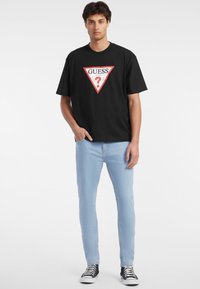 Musta T-paita, jossa on punavalkoinen kolmionmuotoinen logo ja "GUESS?"-teksti, yhdistetty vaaleansinisiin farkkuihin ja mustavalkoisiin lenkkareihin.