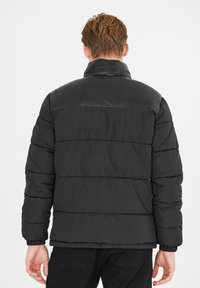 Sort polstret jakke med høj krave, der har syede sektioner og elastiske manchetter. Stoffet har en glat, skinnende overflade.