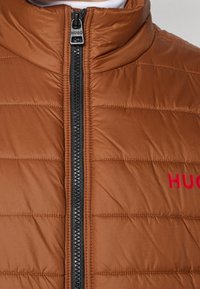 Braune Daunenjacke mit hohem Kragen, vertikaler Steppung, schwarzem Reißverschluss und rotem "HUGO"-Logo auf der Brust.