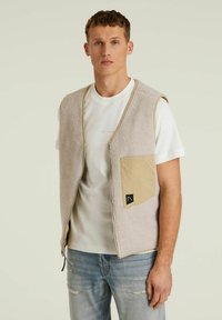 Helebeige fliisvest, millel on lukuga esikülg, tan värvi plaastrikotiga ja musta logoga accent. Selga pandud valge t-särgi peale.