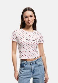 Hvid bomulds cropped T-shirt med et rødt prikket mønster, der har ordet "Strawberry" i sort tekst. Uferdig kant og korte ærmer.