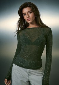 Top verde a maniche lunghe in rete con decorazioni argentate, caratterizzato da uno scollo ampio e una forma aderente, abbinato a jeans grigio chiaro.