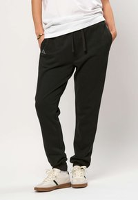 Person som har på seg svart joggebukse med sidelommer, en hvit T-skjorte, hvite joggesko med brune såler og et gullarmbånd på det ene håndleddet.