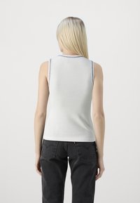 Hvit ermeløs skjorte med rund hals og kontrastsøm, kombinert med svarte jeans. Glatt tekstur, minimal design uten mønstre.
