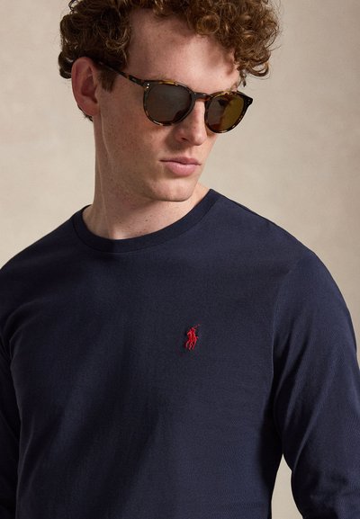 Fiatal férfi göndör hajjal, teknőcpáncél mintás napszemüveget visel, valamint tengerészkék hosszú ujjú inget piros hímzett lovaspóló játékos logóval.