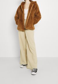 Veste zippée en fausse fourrure marron avec capuche, associée à un pantalon large beige et des baskets noires, créant une tenue décontractée et confortable.