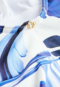 Costum de baie cu bază albă, cu accente albastre, negre și aurii. Include o închizătoare circulară aurie și un material neted, ușor.