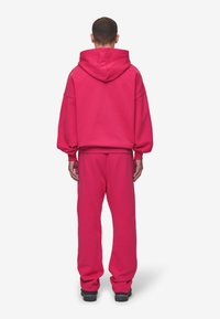 Fleece mikina s kapucňou a jogger nohavicami v jasne ružovej farbe. Mikina má veľkú kapucňu a spustené ramená; nohavice majú elastický pás a manžety.