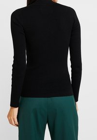 Pull noir côtelé à col roulé avec manches longues, design ajusté, style minimaliste, porté avec un pantalon vert. Texture lisse, sans motifs visibles.