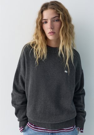 Mladá žena s dlhými vlnitými blond vlasmi, oblečená v tmavosivom svetri cez pruhovanú bordovo-bielu košeľu, na jednoduchom svetlom pozadí.