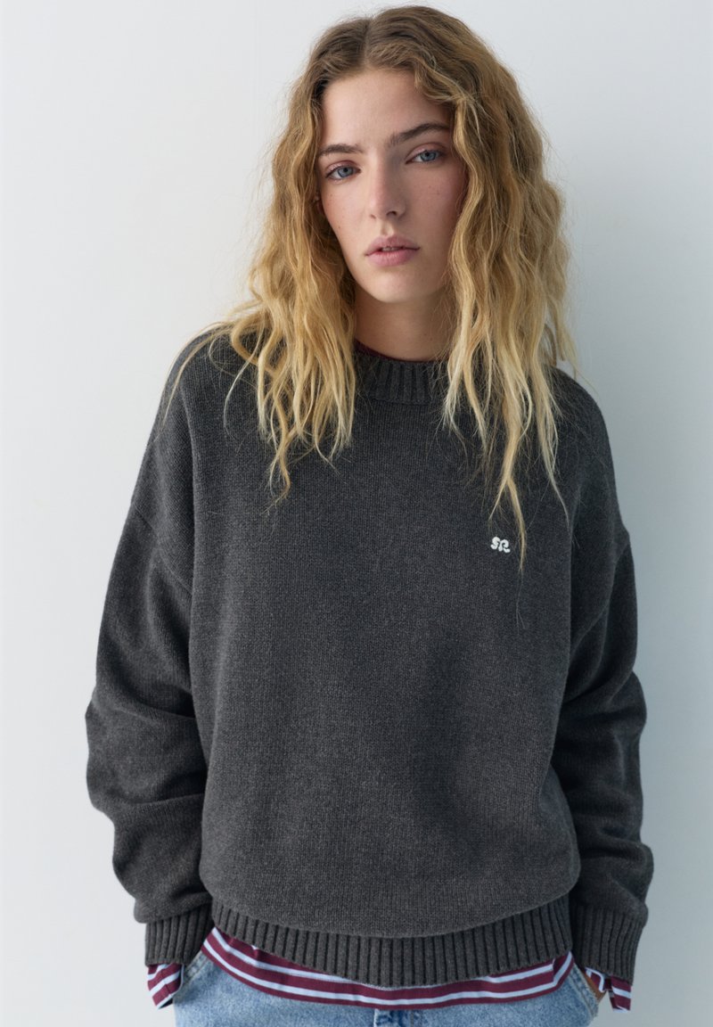 Ung kvinde med langt bølget blondt hår iført en mørkegrå sweater over en stribet, Bordeaux-rød og hvid skjorte mod en ensartet lys baggrund.