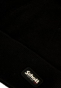 Berretto in maglia nera con texture a coste; presenta un'etichetta rettangolare nera con testo bianco e rosso 'Schott NEW YORK NYC'.