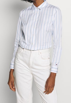 Personne portant une chemise à rayures verticales bleu clair et blanc, rentrée dans un pantalon blanc taille haute, sur un fond uni.