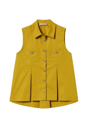 Donna che indossa un top giallo senape senza maniche con bottoni e gonna abbinata, accessoriata con collana e bracciali d'oro, guarda a sinistra.