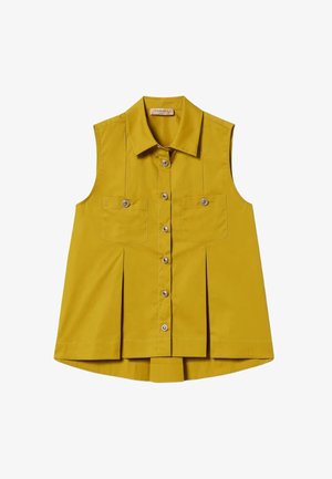 Donna che indossa un top giallo senape senza maniche con bottoni e gonna abbinata, accessoriata con collana e bracciali d'oro, guarda a sinistra.