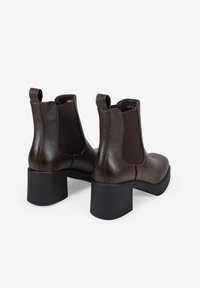 Bottines en cuir marron avec panneaux latéraux élastiques, bout arrondi et talons épais noirs. Texture lisse avec des détails de design minimalistes.