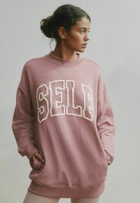 Roosa oversized lõikega dressipluus soonikkoes kaeluse ja mansettidega, mille esiküljel on valge tekstuurse tikkimisega sõna "SELF".