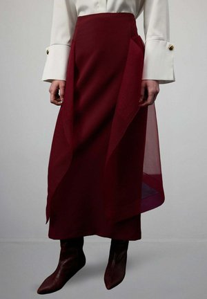 Kobieta w bordowej spódnicy z wysokim stanem z asymetryczną przezroczystą narzutką oraz białą bluzką z szerokimi mankietami i złotymi guzikami.