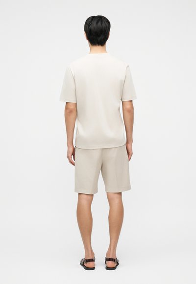 Άνδρας που στέκεται με την πλάτη, φοράει μπεζ κοντομάνικο πουκάμισο, ταιριαστά σορτς και μαύρα σανδάλια μπροστά σε απλό λευκό φόντο.