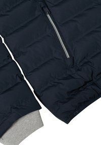 Veste matelassée bleu marine avec une fermeture éclair argentée, présentant un poignet gris et une finition brillante. Montre un tissu texturé sur certaines sections des manches.