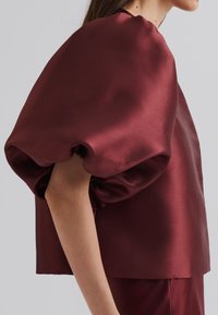 Haut en satin bordeaux avec des manches bouffantes volumineuses, coupe carrée, texture lisse et encolure ronde, porté sur une jupe assortie.