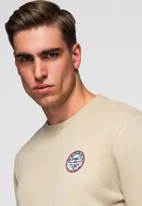 Beige sweatshirt met een ronde logopatch met blauwe en rode accenten. De stof oogt glad met een ronde halslijn en geribde manchetten.