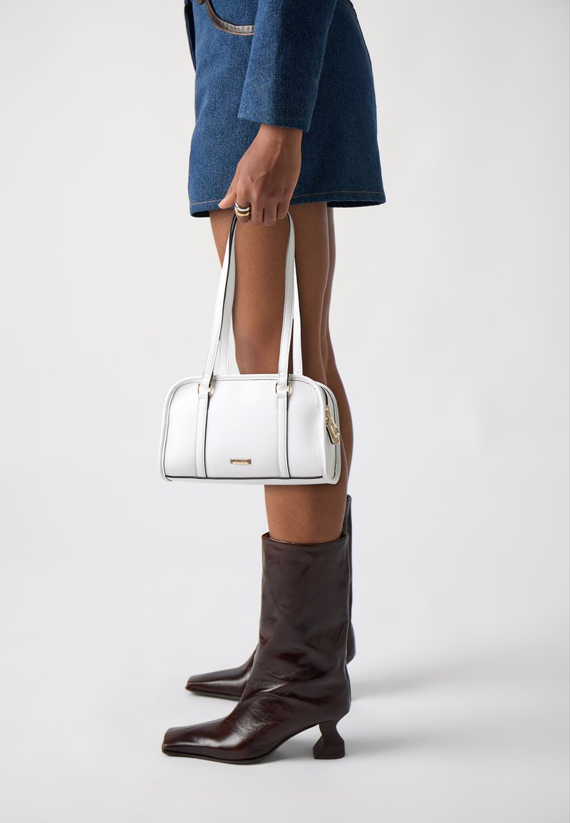 Sac à main en cuir blanc avec deux anses et quincaillerie dorée, affichant un design structuré et des coutures noires contrastantes. Bottes marron jusqu'au genou.