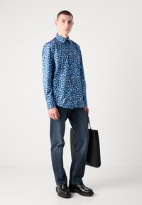 Chemise à boutons bleue à motifs floraux avec manches longues, associée à un jean en denim foncé et des chaussures noires, tenant un tote bag noir.