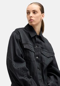 Czarna kurtka jeansowa z klasycznym kołnierzem, dwoma kieszeniami na piersi oraz ćwiekami na lewym rękawie. Tradycyjne zapinanie na guziki z przodu.