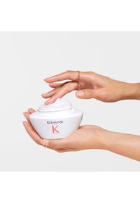 Witte keramische pot met een ronde vorm, met Kérastase branding in het rood. Crèmige product is iets zichtbaar bovenaan; vastgehouden door een hand.