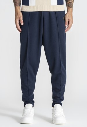 Uomo che indossa pantaloni blu navy a cavallo basso con gamba affusolata e sneakers bianche, in piedi davanti a uno sfondo uniforme.