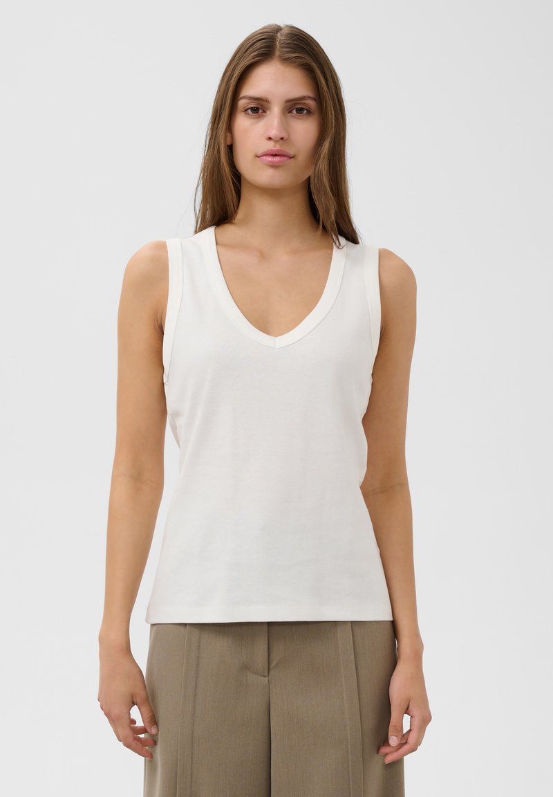 Jeune femme aux longs cheveux bruns portant un haut blanc sans manches à col en V et un pantalon beige, debout devant un fond clair uni.