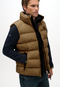 Gilet imbottito color tan con colletto alto, chiusura con zip e tasche laterali, indossato sopra un maglione blu scuro a trecce; presenta un patch con logo.