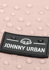 Čierna gumená nálepka s bielym textom "JOHNNY URBAN" na textúrovanej svetloružovej hladine so zdvihnutými vzormi kvapiek vody.