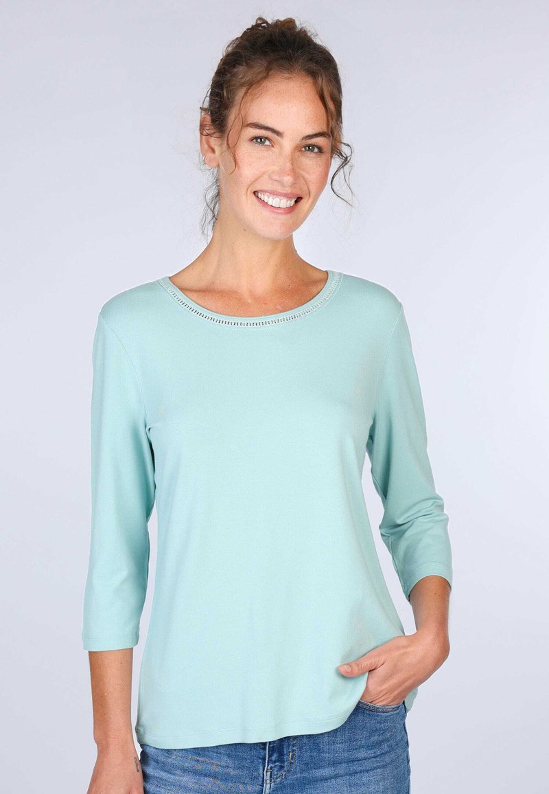 Hellblaues Oberteil mit drei Viertelärmeln und rundem Ausschnitt, glatter Textur und dekorativen Nähten entlang des Ausschnitts.