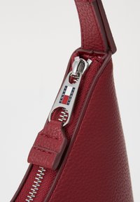 Rode leren handtas met textuur, voorzien van een zilveren rits trekker, gemarkeerd met "TOMMY JEANS," en een gladde leren ritslip.