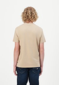 Beige bomull t-skjorte med rund hals og korte ermer, vist fra baksiden. Har en glatt tekstur og er kombinert med mørke denimbukser.